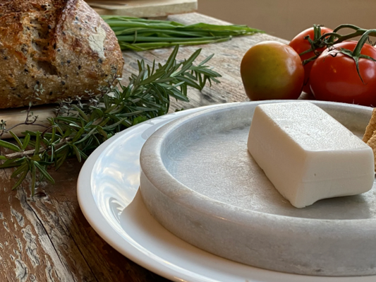 Block of Let Them Eat Vegan Mozzarella on a stone platter with bread, tomatoes, and herbs on a wooden table.