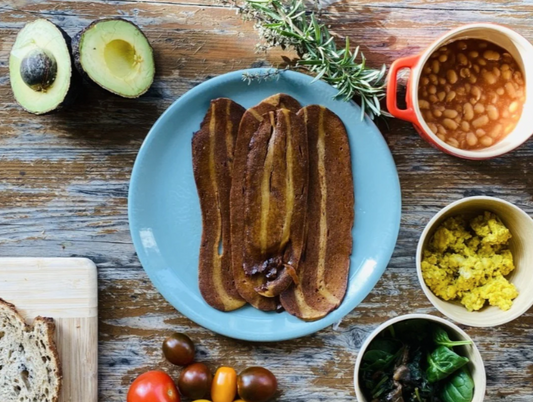 Let Them Eat Vegan NOT Bacon Slices on a blue plate with avocado, beans, and other ingredients on a wooden table