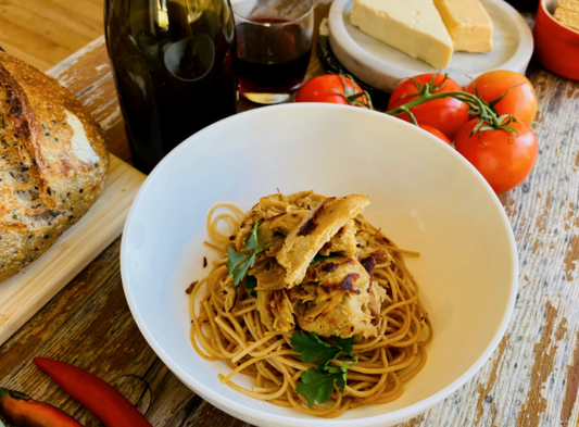 Spaghetti dish with Let Them Eat Vegan NOT Chook and herbs in a white bowl on a wooden table with bread, tomatoes, and a bottle.