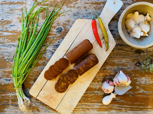 Wooden cutting board with sliced Let Them Eat Vegan NOT Pepperoni, green onions, red and green peppers, and garlic cloves on a rustic wooden surface.