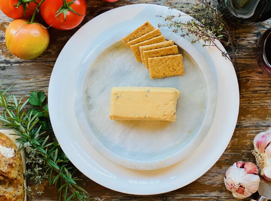 Block of Let Them Eat Vegan Onion & Chive Cheddar Block on a marble plate with crackers and tomatoes on a wooden table