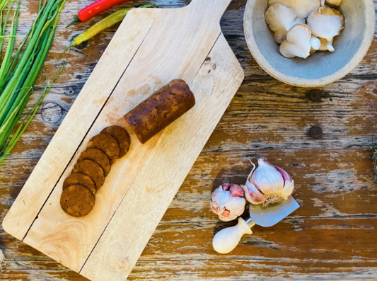 Wooden cutting board with sliced Let Them Eat Vegan NOT Salami, garlic, and a bowl of mushrooms on a rustic wooden table.