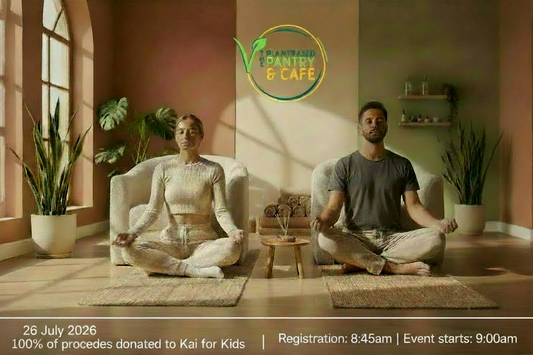 A man and a woman meditate cross-legged in matching beige outfits inside a sunlit room with terracotta and olive walls, potted plants, and woven mats.
