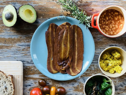 Let Them Eat Vegan NOT Bacon Slices on a blue plate with avocado, beans, and other ingredients on a wooden table