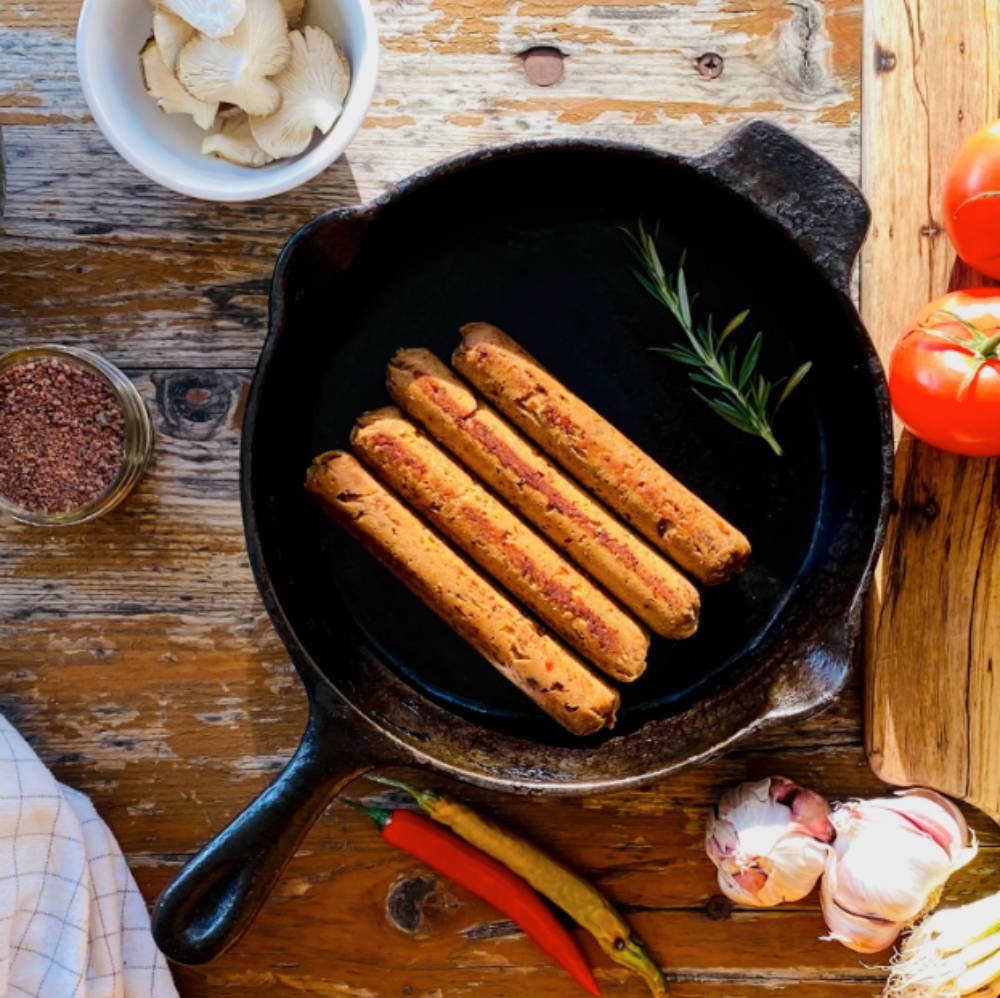 Let Them Eat Vegan NOT Chorizo sausages cooking in a cast iron skillet on a wooden table with vegetables.