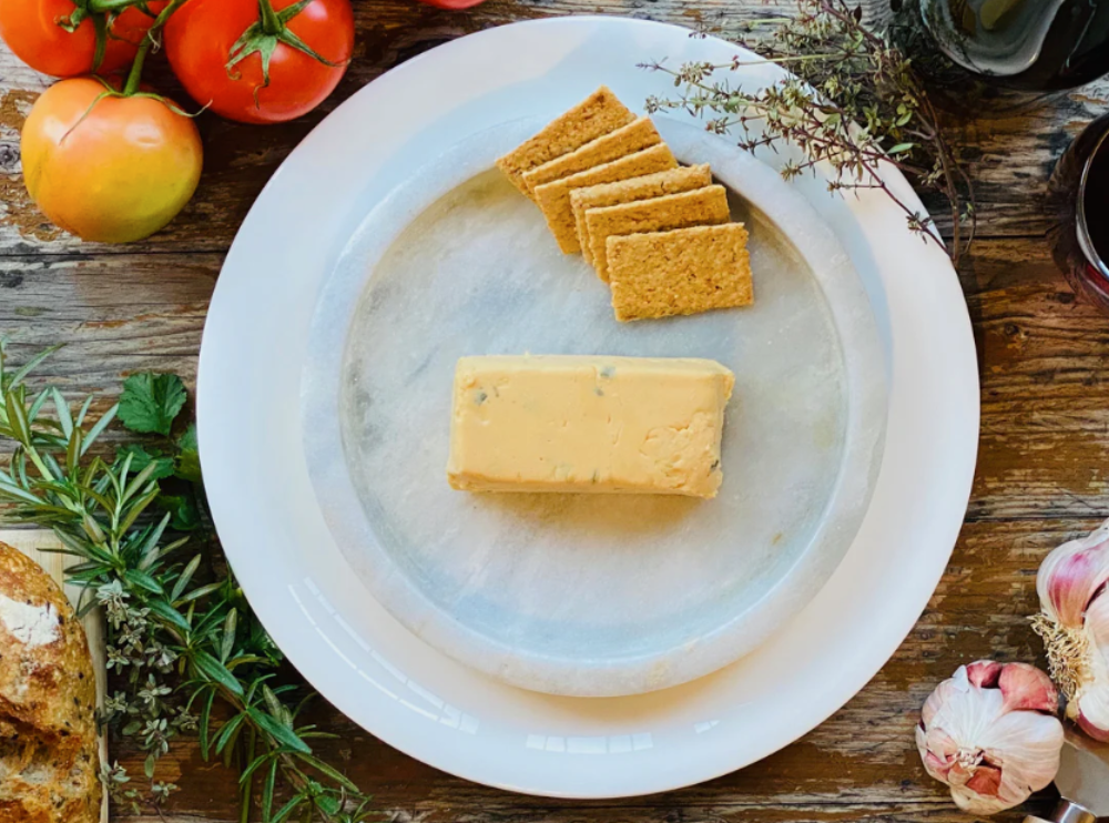 Block of Let Them Eat Vegan Onion & Chive Cheddar Block on a marble plate with crackers and tomatoes on a wooden table