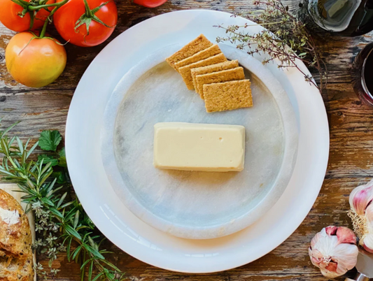 Let Them Eat Vegan Smoked Gouda Block on a marble slab with crackers and tomatoes on a wooden surface