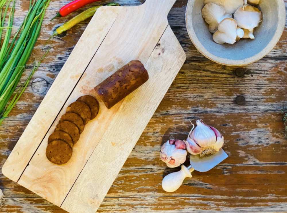 Wooden cutting board with sliced Let Them Eat Vegan NOT Salami, garlic, and a bowl of mushrooms on a rustic wooden table.