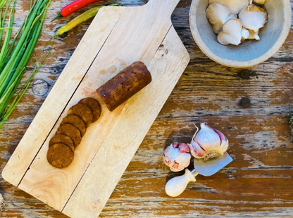 Wooden cutting board with sliced Let Them Eat Vegan NOT Salami, garlic, and a bowl of mushrooms on a rustic wooden table.
