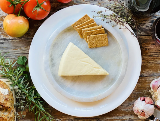 Let Them Eat Vegan Camembert Wedge on a marble platter with crackers, surrounded by fresh ingredients on a wooden table.