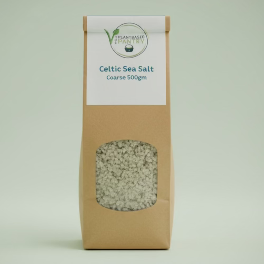A 500g bag of PlantBased Pantry Celtic Sea Salt, Coarse, standing on a wooden table in a store setting. The bag is made of brown kraft paper with a clear window showing the coarse salt inside and a white label at the top.