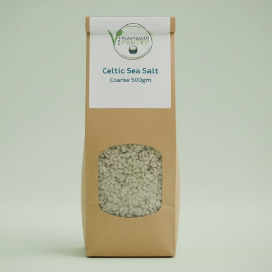 A 500g bag of PlantBased Pantry Celtic Sea Salt, Coarse, standing on a wooden table in a store setting. The bag is made of brown kraft paper with a clear window showing the coarse salt inside and a white label at the top.