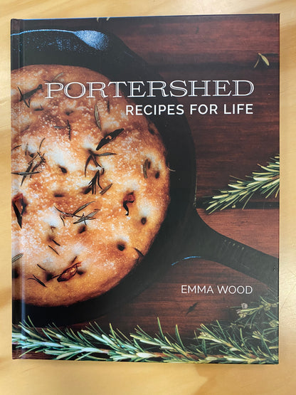 Portershed Recipes for Life cookbook by Emma Wood, featuring a rustic baked bread in a cast iron skillet on a dark wooden background, placed on a light wood surface.