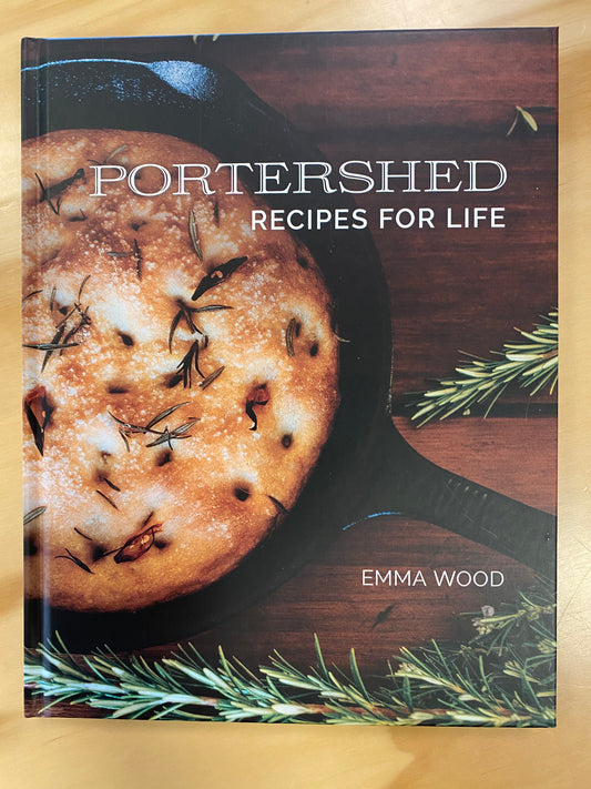 Portershed Recipes for Life cookbook by Emma Wood, featuring a rustic baked bread in a cast iron skillet on a dark wooden background, placed on a light wood surface.