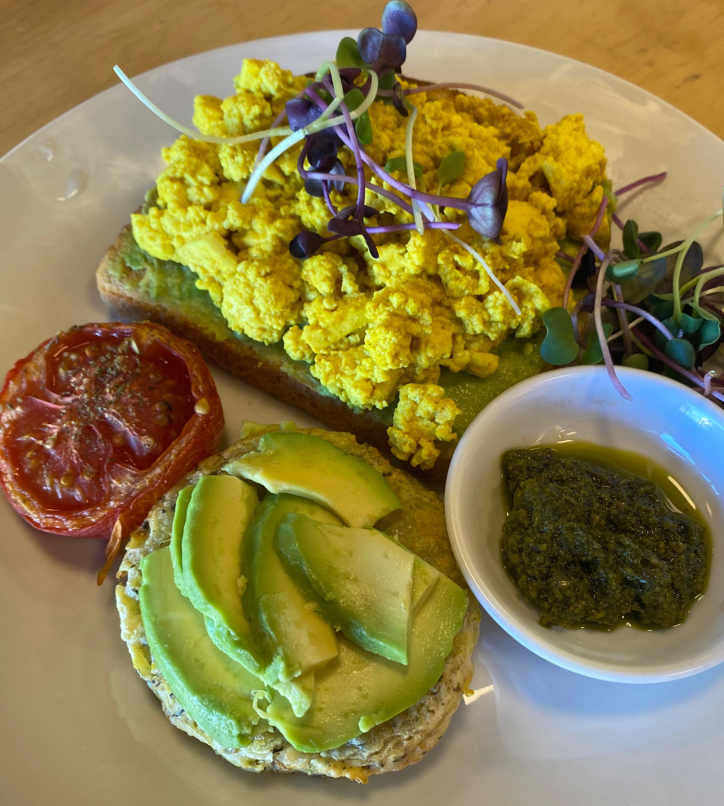 Plate of PBCafe Tofu Scramble with , scrambled tofu on toast, avocado on a potato rosti, roast tomato and a side of fresh basil pesto on a wooden table.