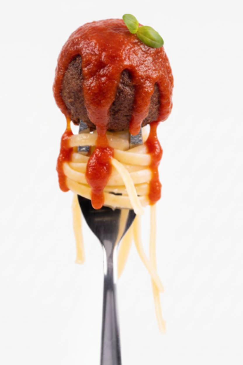 A close-up of a fork holding a meatball topped with a small green garnish and pasta on a white background.
