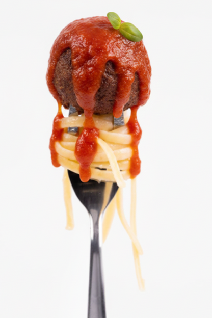 A close-up of a fork holding a meatball topped with a small green garnish and pasta on a white background.
