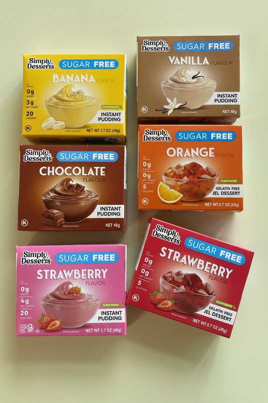 Six boxes of Simply Desserts sugar-free instant pudding in assorted flavors arranged on a light surface: Banana, Vanilla, Chocolate, Orange, Strawberry, and another Strawberry box. The packaging colors differ by flavor, with yellow, brown, dark brown, orange, pink, and red boxes.